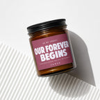 Amber jar candle with dark lid and maroon label reading "OUR FOREVER BEGINS".