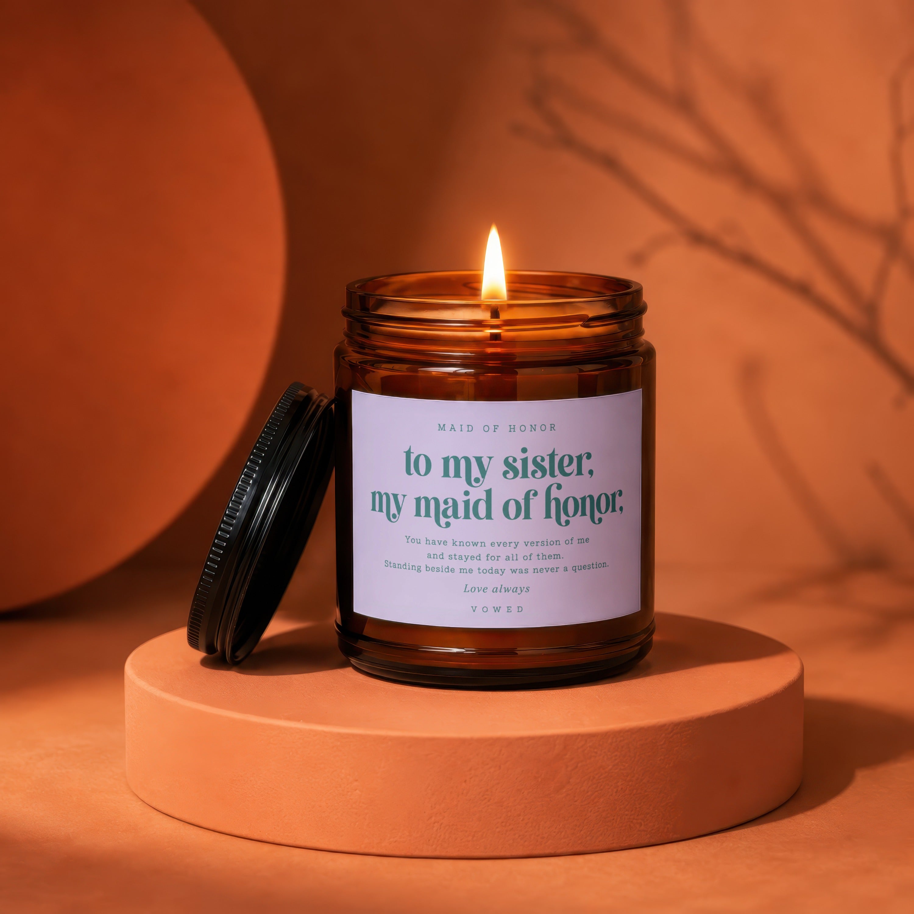 A lit candle in a amber glass jar with a label reading "Maid of Honor to my sister".