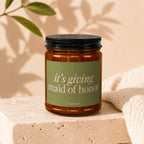Amber glass candle with a green label reading "it's giving maid of honor".