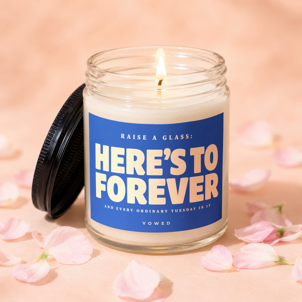 Glass candle with lit wick and blue label reading "HERE'S TO FOREVER".