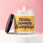 Lit candle in glass jar with yellow label that reads "Mission Impossible Accomplished"