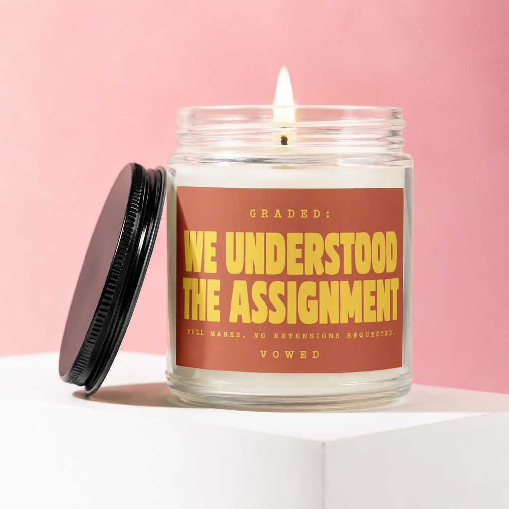Lit candle in a jar with a black lid and label that reads "GRADED: WE UNDERSTOOD THE ASSIGNMENT".