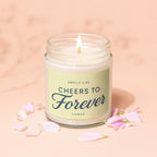 Jar candle with lit flame, reads "Smells like Cheers to Forever", surrounded by petals.