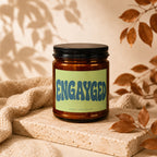 Brown glass candle with "ENGAYGED | VOWED | HAND POURED" on a light green label.