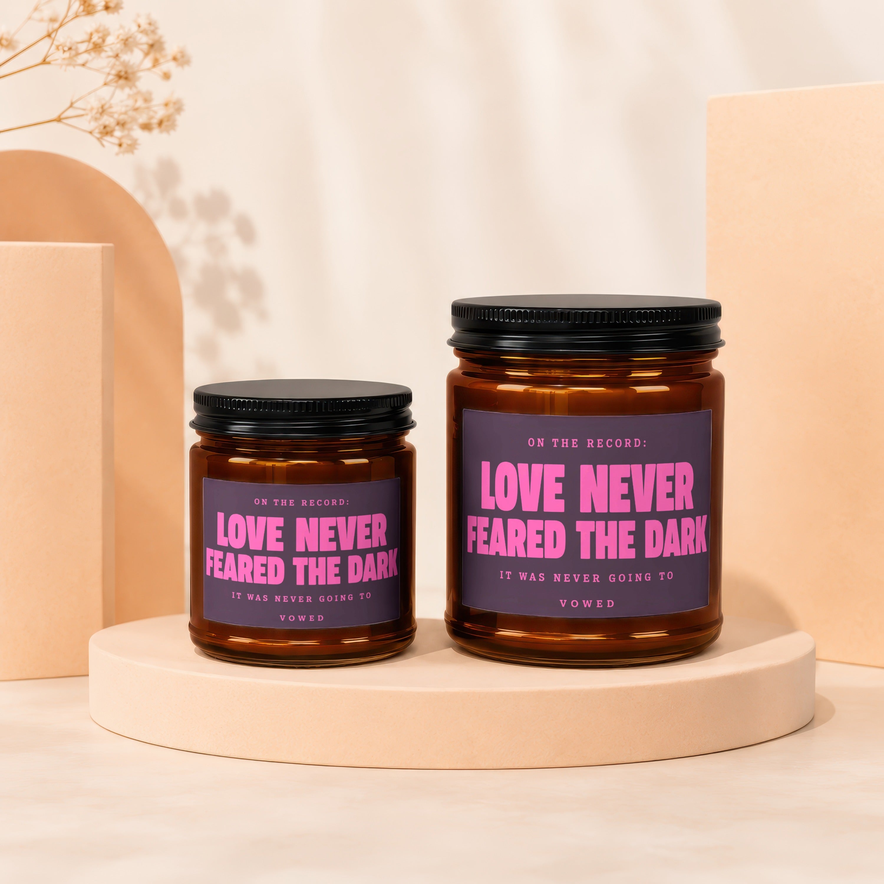 Two amber glass candles with black lids both read "Love Never Feared The Dark".
