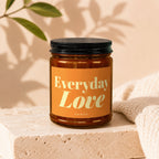 Amber glass candle labeled "Everyday Love" by Vowed on a stone block with a beige knit fabric.