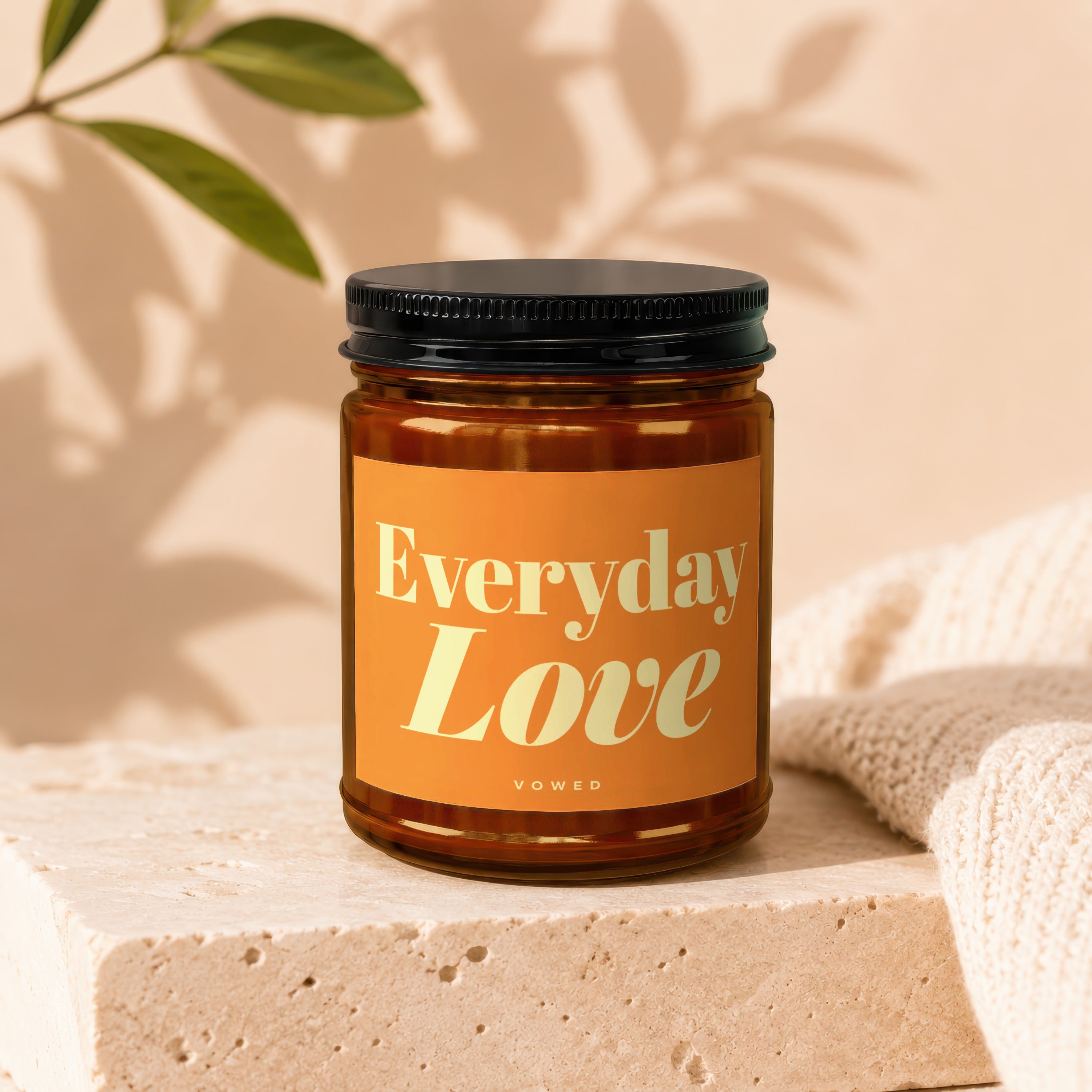 Amber glass candle labeled "Everyday Love" by Vowed on a stone block with a beige knit fabric.