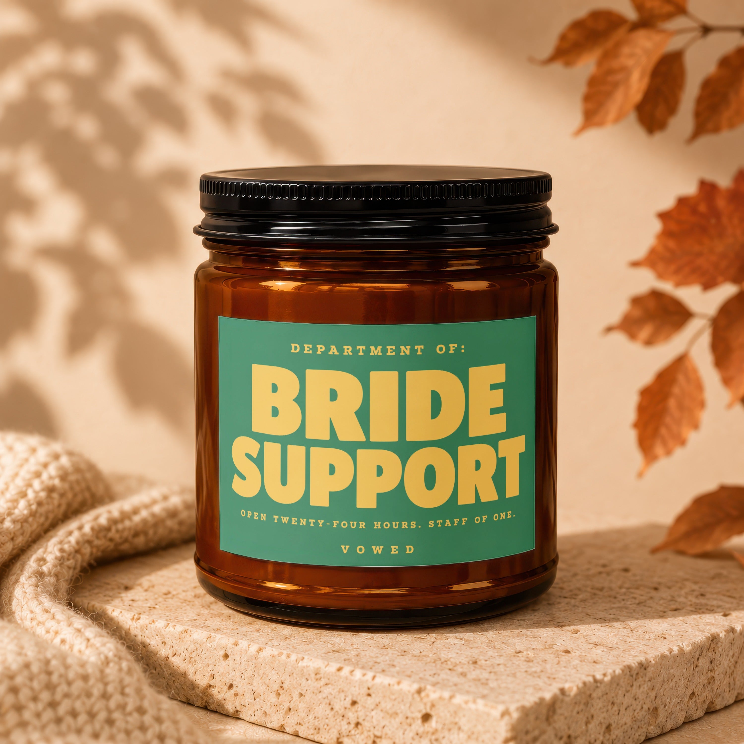 Brown jar candle with green label reading "DEPARTMENT OF: BRIDE SUPPORT OPEN TWENTY-FOUR HOURS. STAFF OF ONE. VOWED".