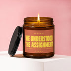 Amber candle with lit wick and black lid, labeled "We Understood The Assignment".