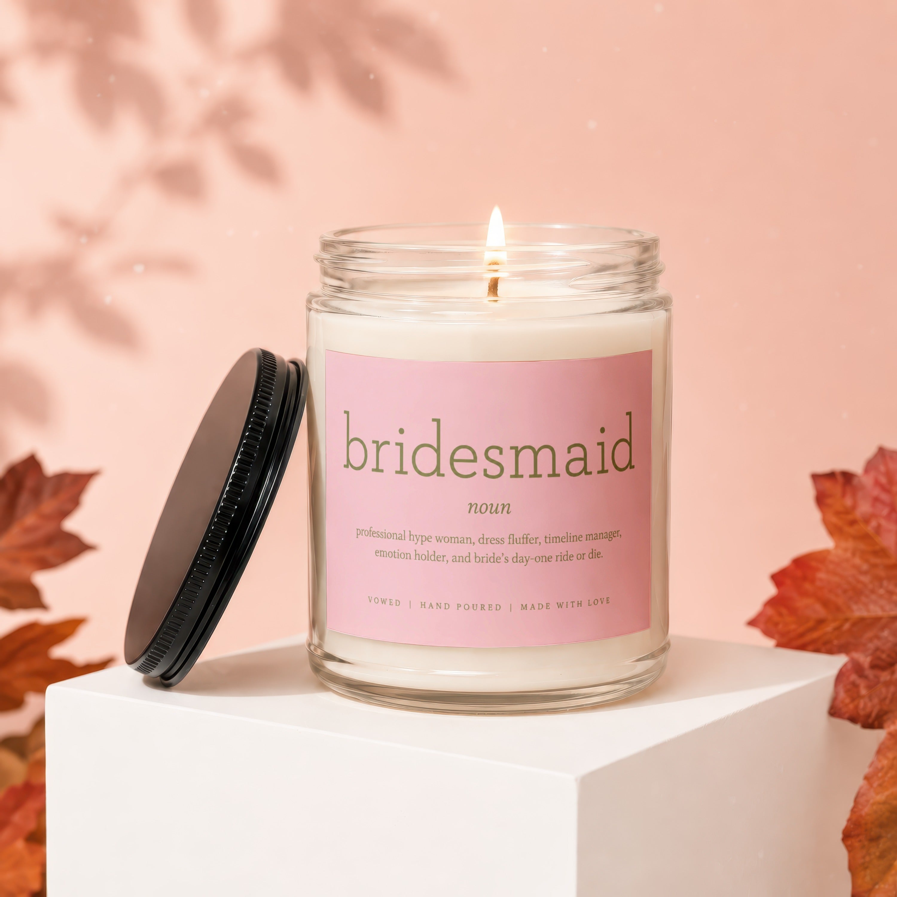 Glass candle with "bridesmaid" text, lid nearby, on display riser.