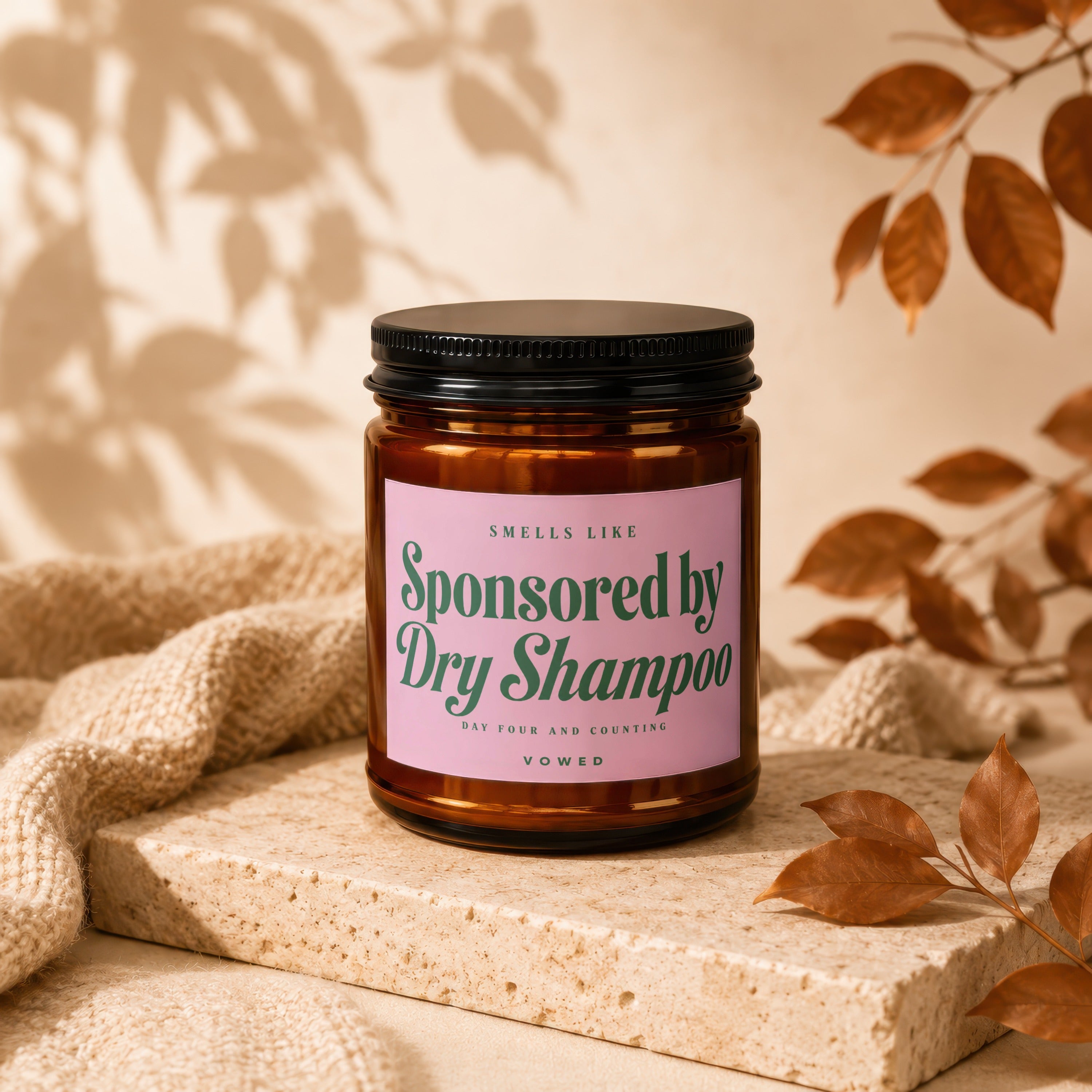 Candle labeled "Sponsored by Dry Shampoo" on a stone stand with textured fabric and leaves.