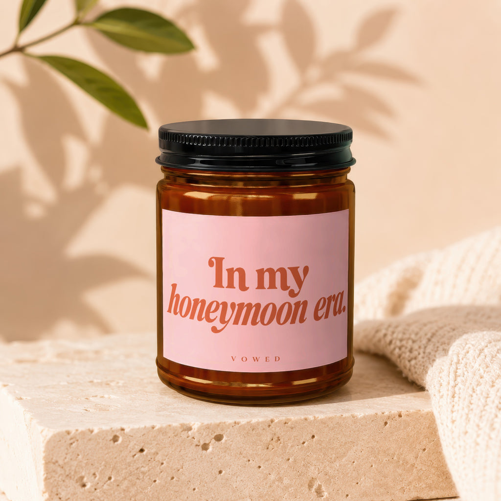 Brown glass candle with pink label reading "In my honeymoon era. VOWED."