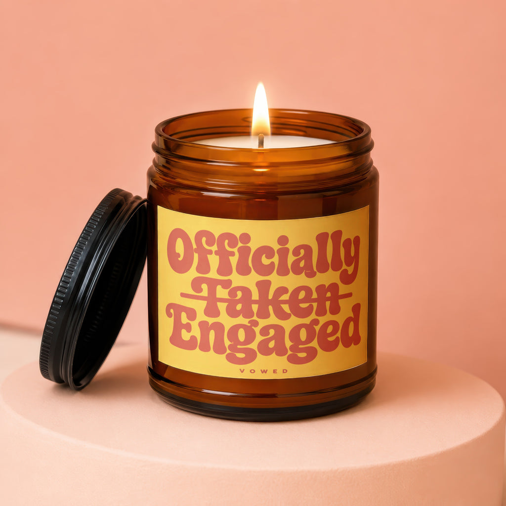 Lit brown glass candle with a yellow label reading "Officially Taken Engaged".