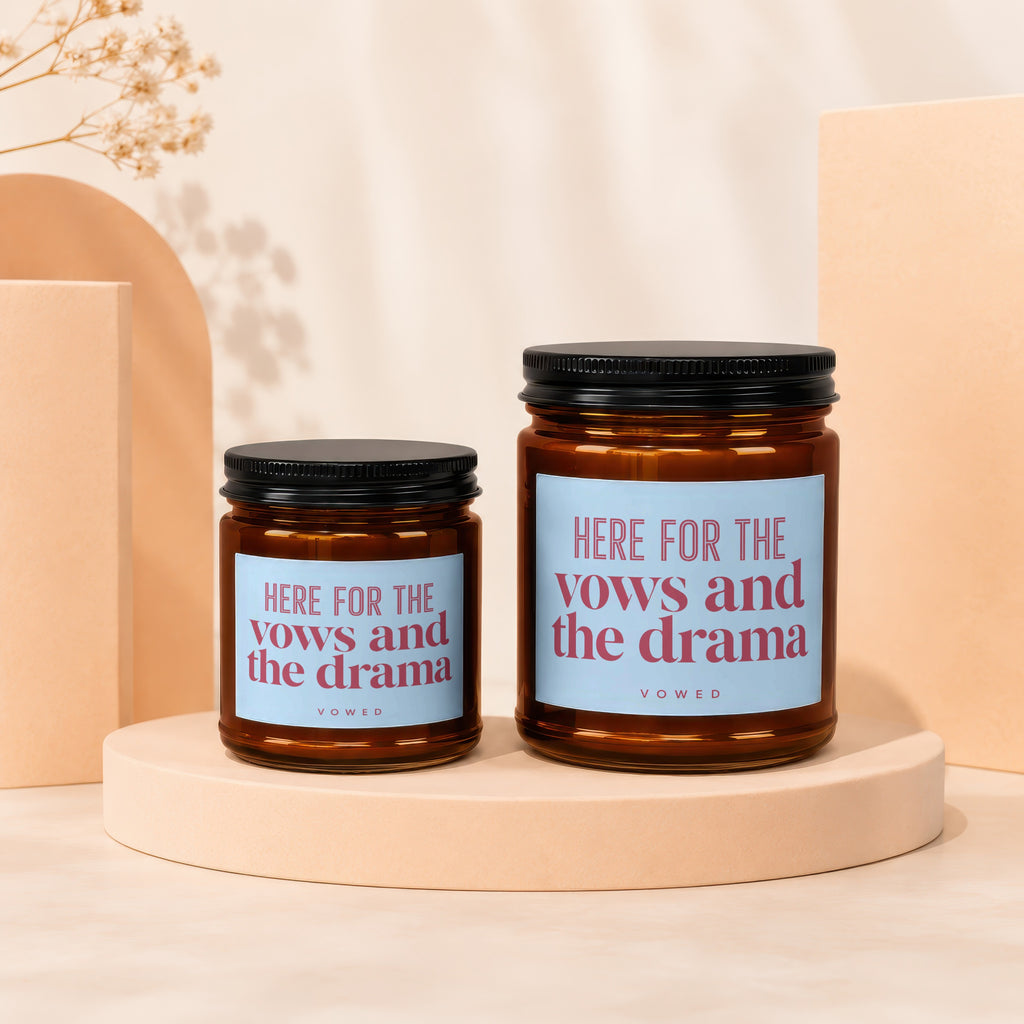 Two amber glass candles with black lids and blue labels reading "HERE FOR THE vows and the drama."