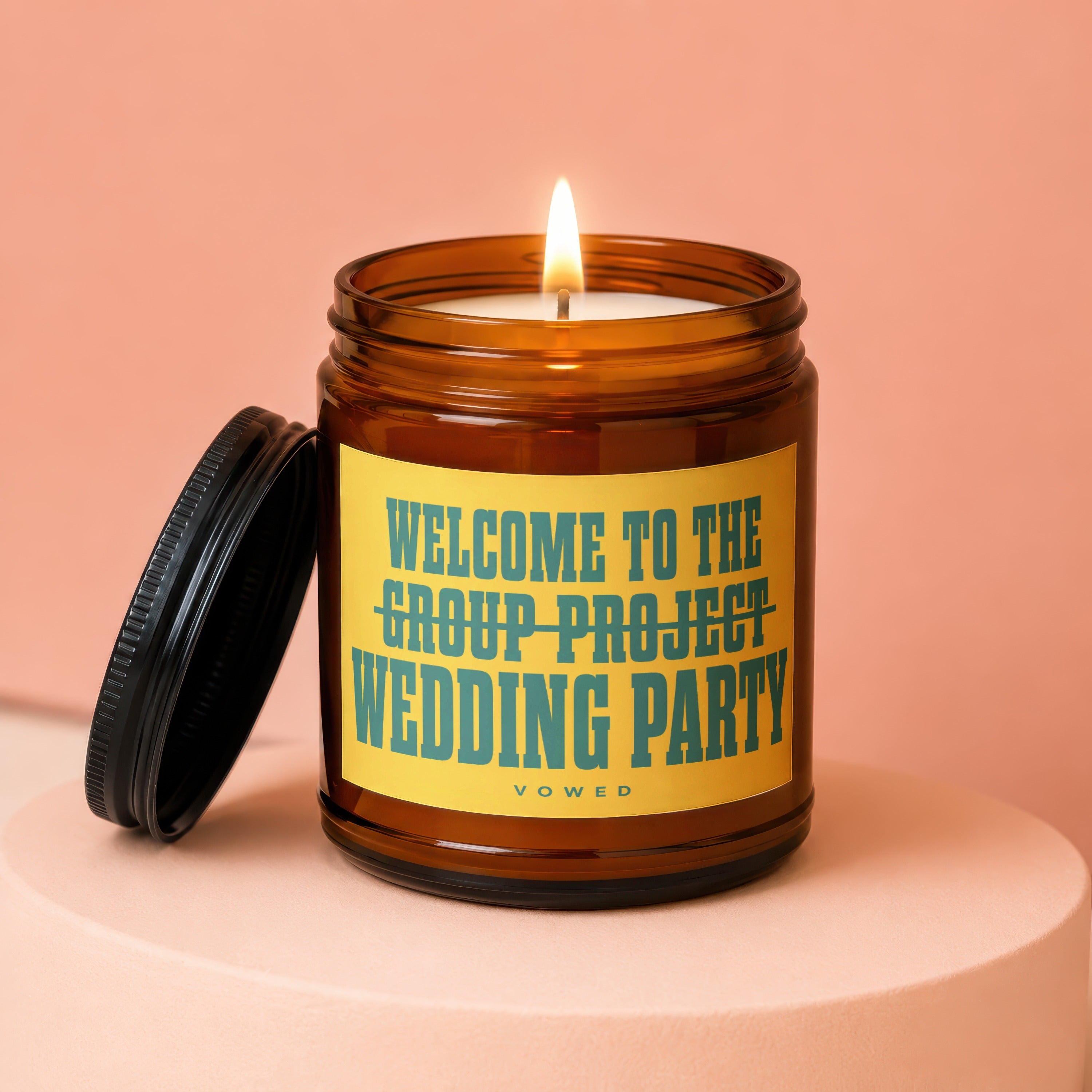 Lit amber jar candle with a yellow label reading "WELCOME TO THE GROUP PROJECT WEDDING PARTY".
