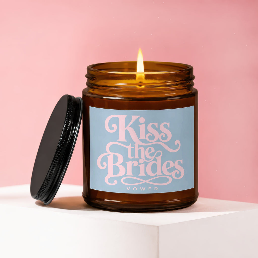 Amber jar candle with lit flame, blue label reads "Kiss the Brides VOWED".
