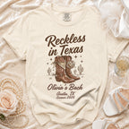Cream t-shirt with "Reckless in Texas" and cowboy boots graphic, "Olivia's Bach Austin, TX Summer 2026".