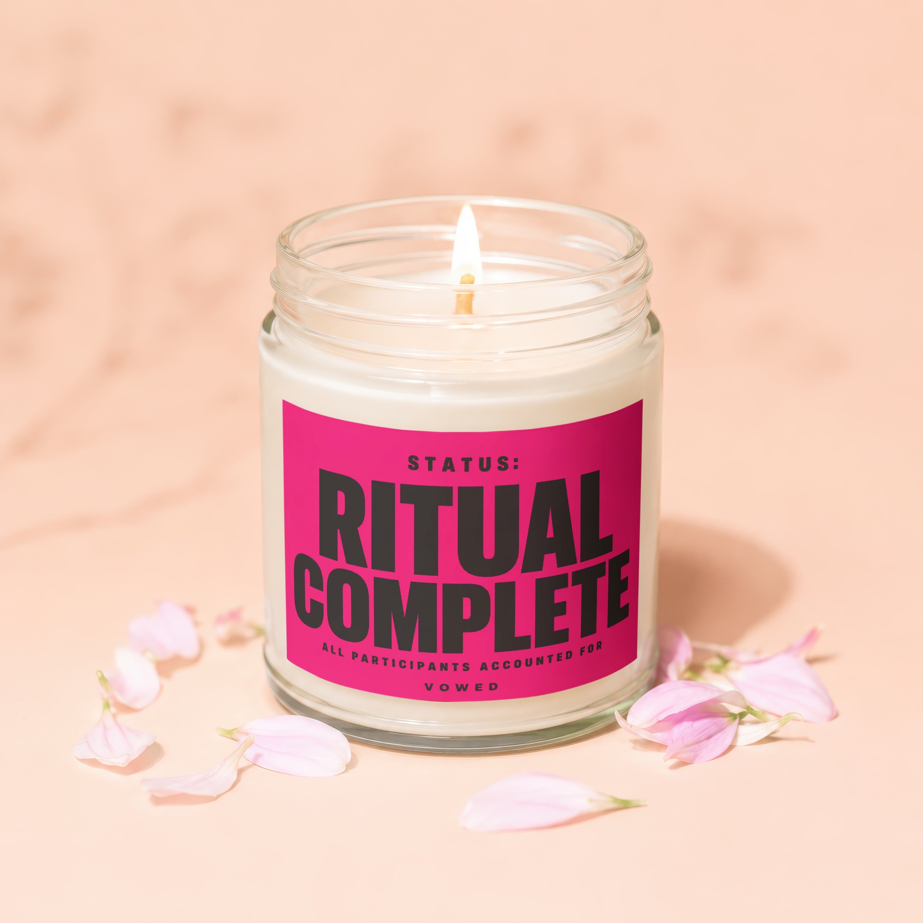 Glass jar candle with pink label reading "STATUS: RITUAL COMPLETE" surrounded by petals.