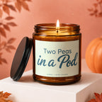 Amber candle with "Two Peas in a Pod" text, lid beside it, pumpkin and leaves.