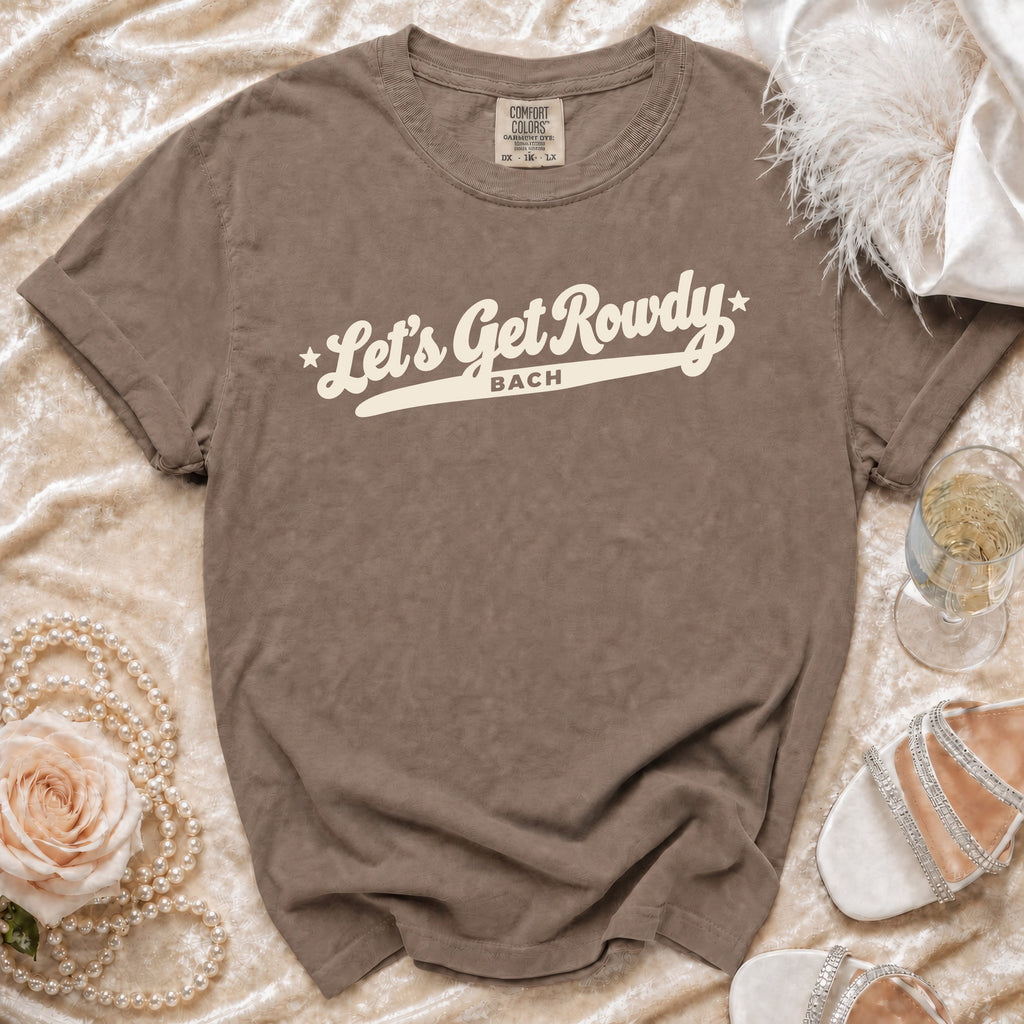 A brown graphic t-shirt with the white text Let's Get Rowdy Bach, displayed on a satin background with a rose, pearls, and heels.