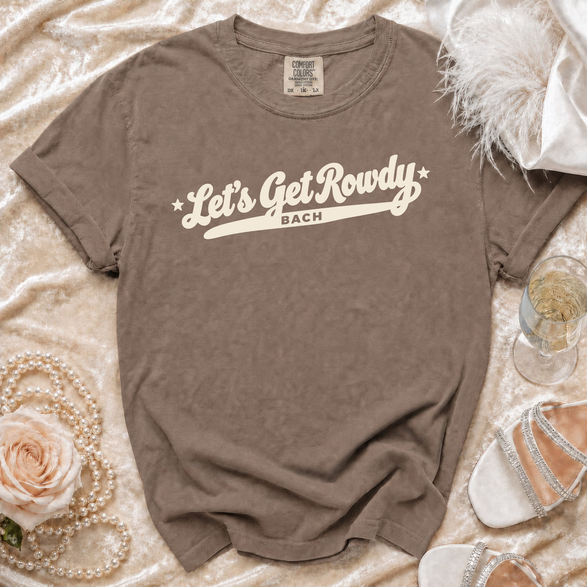 A brown graphic t-shirt with the white text Let's Get Rowdy Bach, displayed on a satin background with a rose, pearls, and heels.