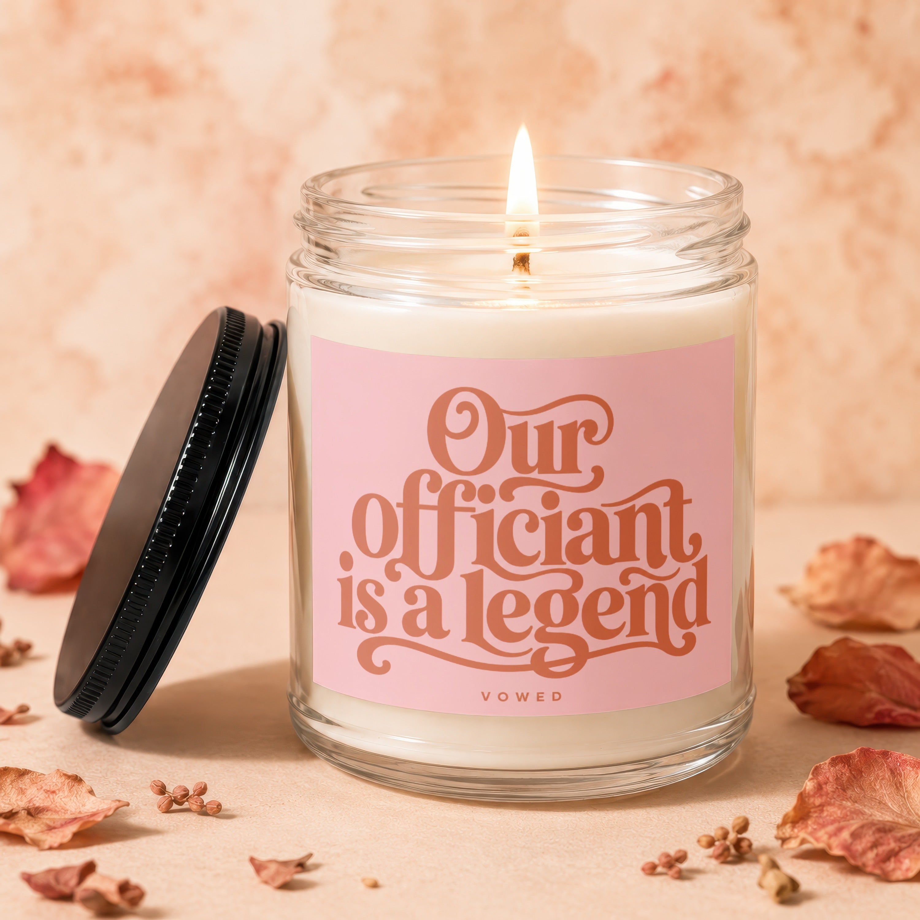 Jar candle with lit wick, pink label reading "Our officiant is a legend"