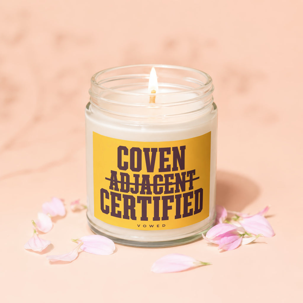 Candle with "Coven Adjacent Certified" text on yellow label, surrounded by pink petals.