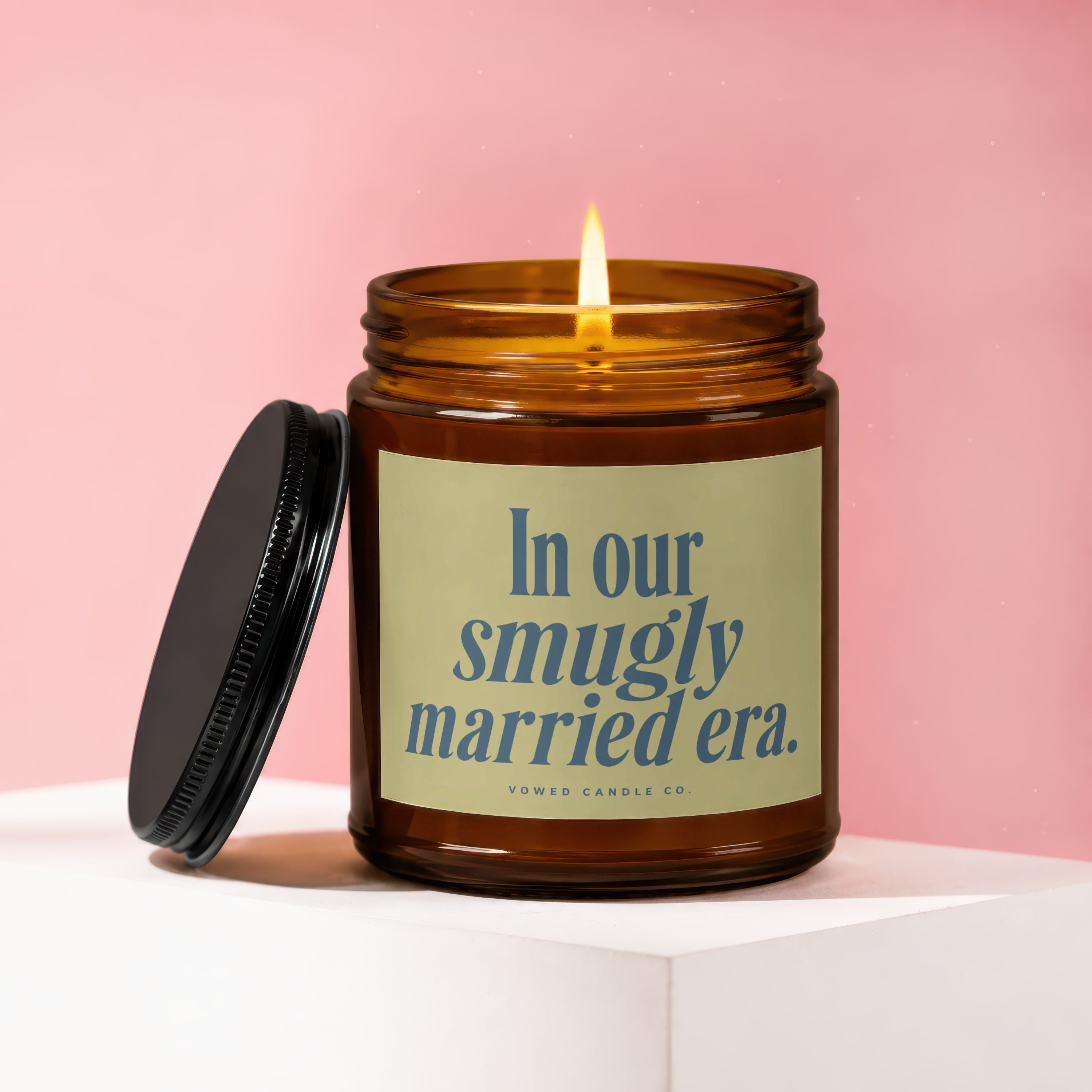 Amber candle with lit flame and black lid, displaying "In our smugly married era."