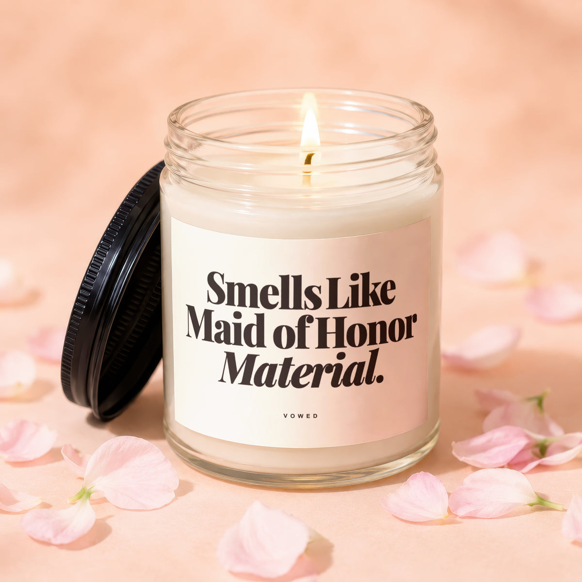 Candle with "Smells Like Maid of Honor Material." text, lid off, surrounded by petals.