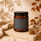 Amber candle with black lid, label reads "The Spark Before Forever".