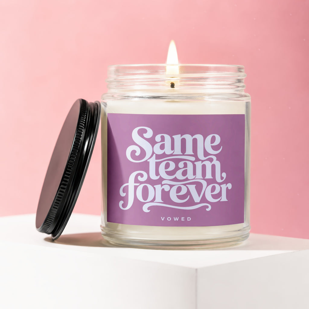 Glass candle with lit wick and "Same team forever vowed" on label.