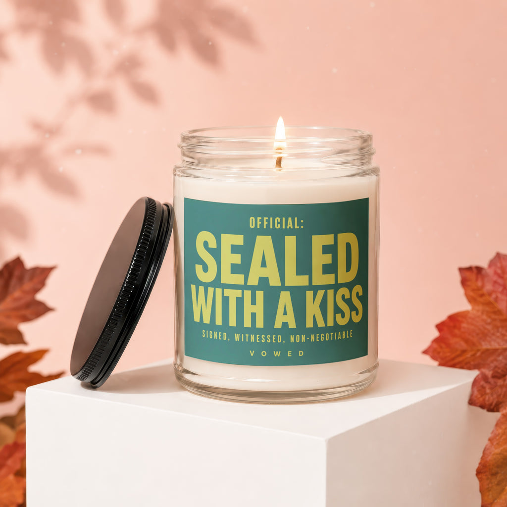 Glass candle with lit wick and black lid, text says "OFFICIAL: SEALED WITH A KISS VOWED"