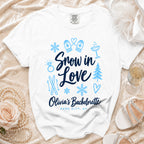 A white t-shirt features blue text reading Snow in Love, Olivia's Bachelorette, Park City, UT, surrounded by winter and bachelorette graphics.
