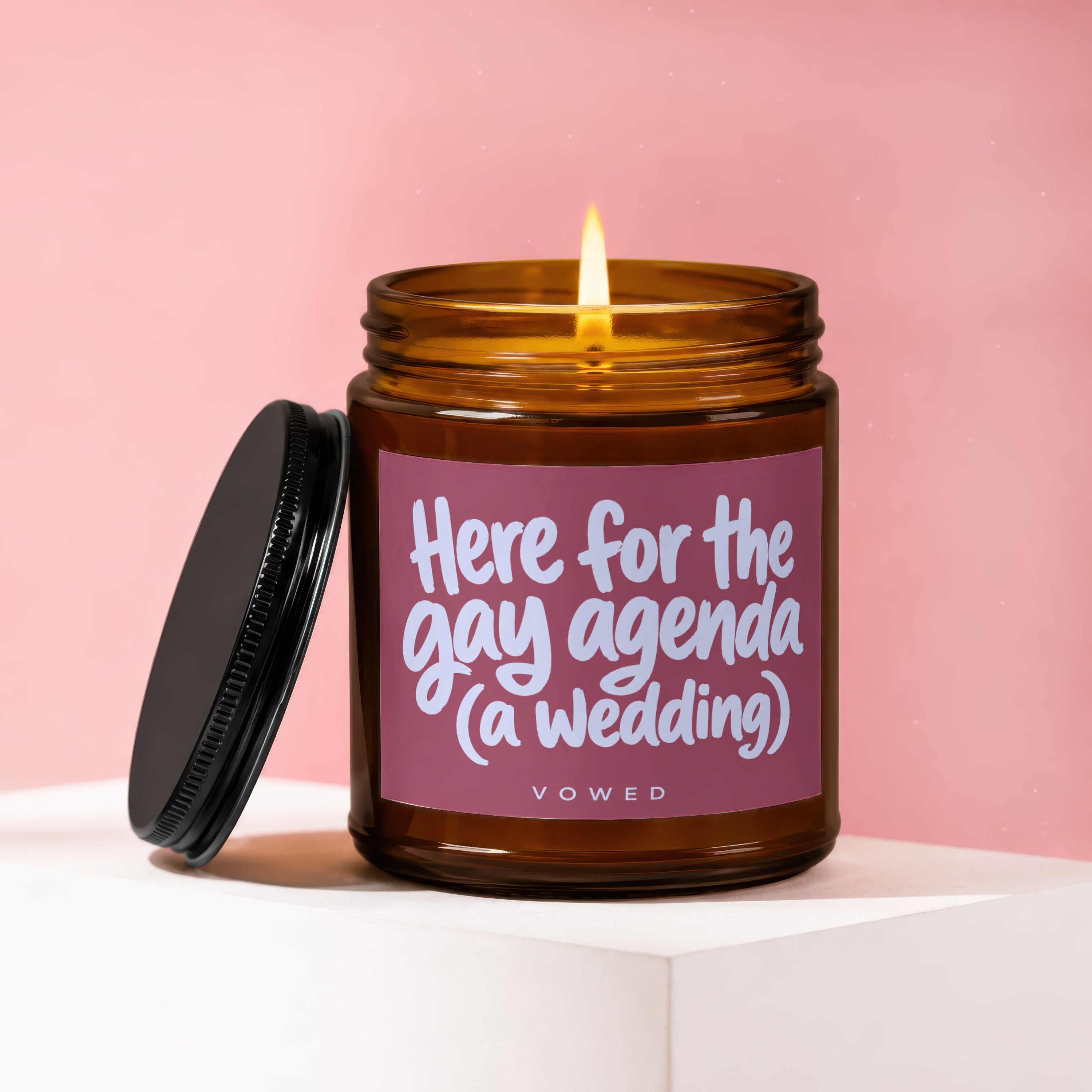 Amber candle with lit flame on pink background, with text "Here for the gay agenda (a wedding)".