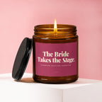 Amber glass candle with lit wick, pink label reads "The Bride Takes the Stage. Standing Ovation Expected."