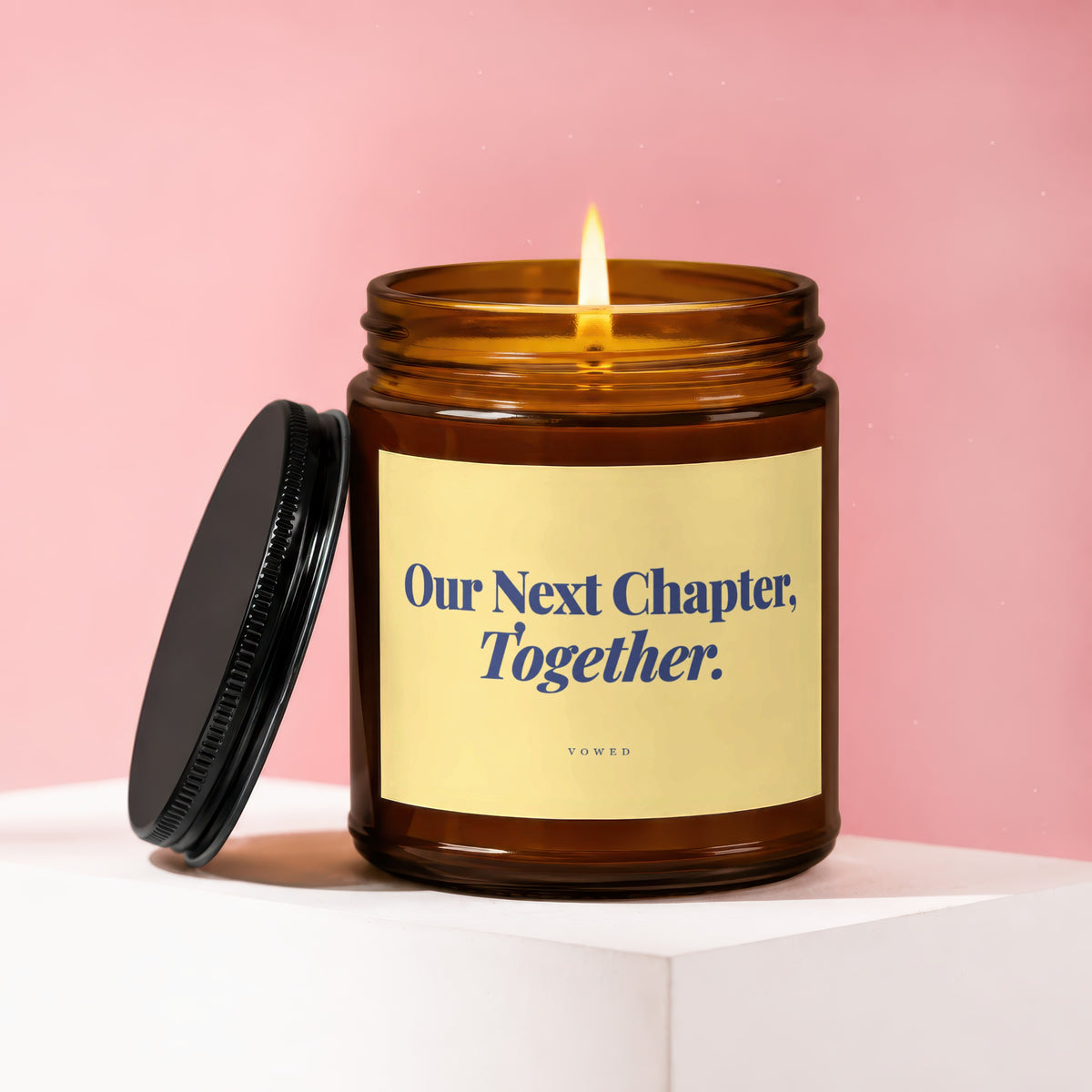 Amber glass candle with a lit wick and black lid, reads "Our Next Chapter. Together."