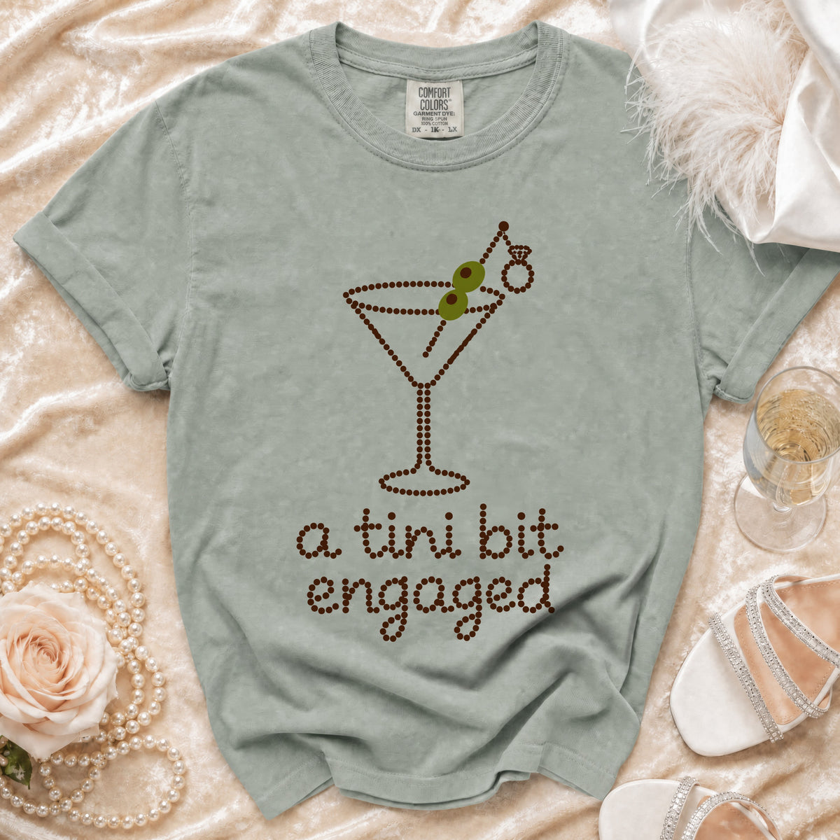 Sage green t-shirt featuring a beaded martini glass graphic with the text a tini bit engaged.