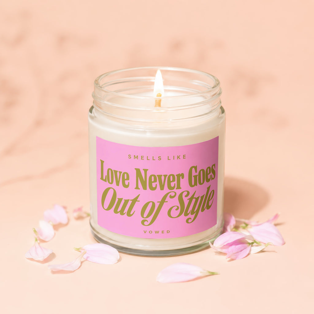 Glass jar candle with lit wick featuring pink label that reads "Smells Like Love Never Goes Out of Style."
