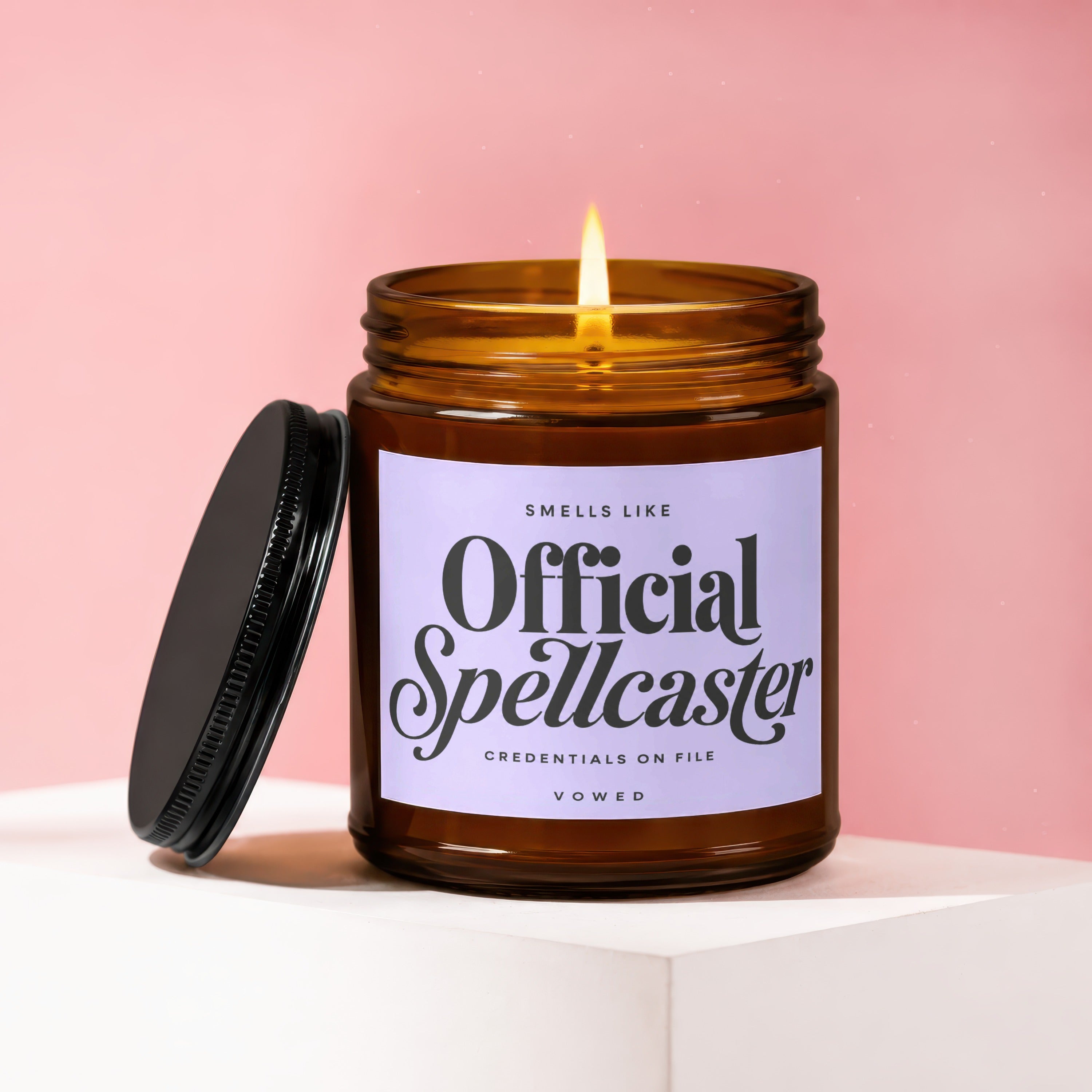 Amber jar candle with lit wick and black lid, displays "Smells like Official Spellcaster" text.