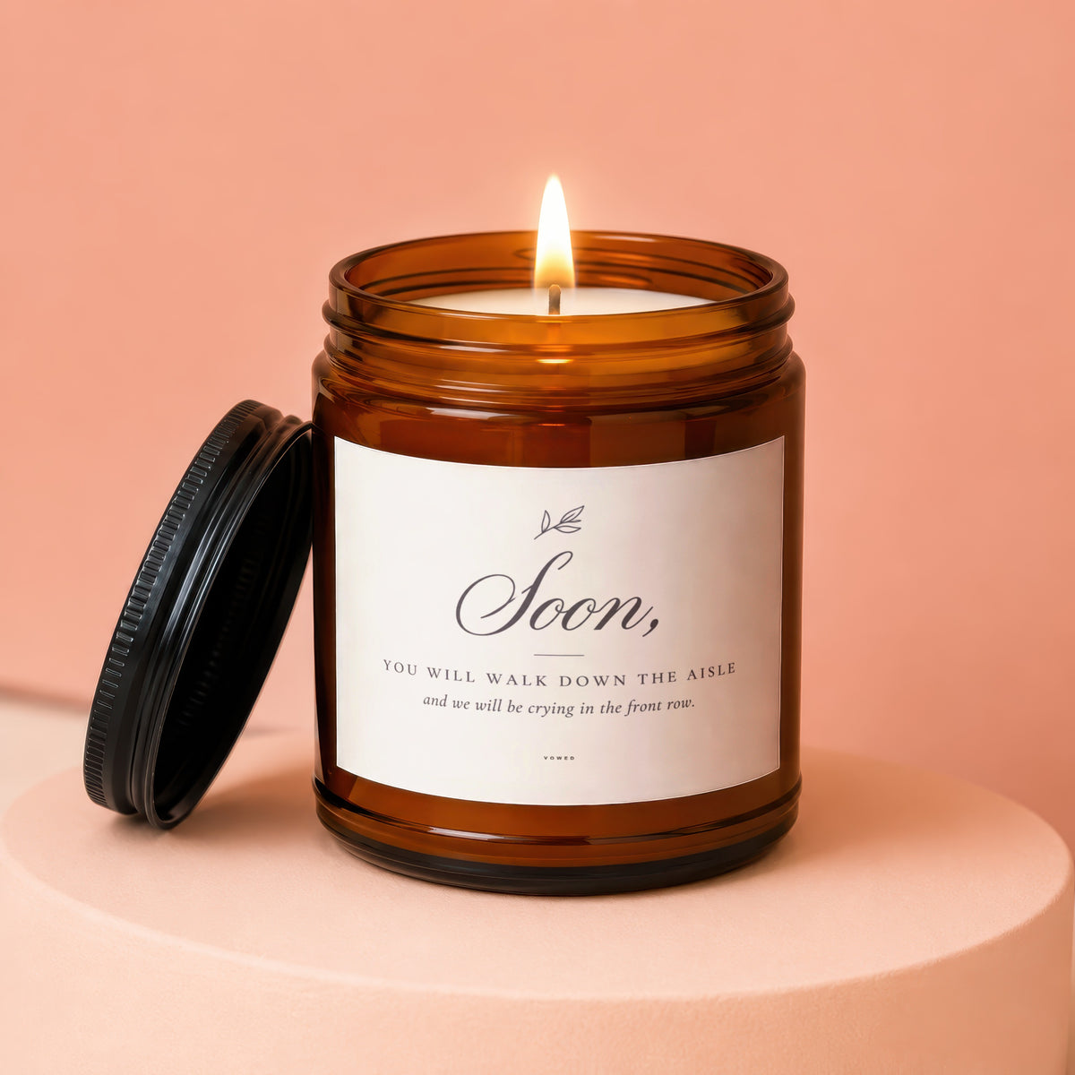 Amber jar candle with lit flame and lid. Label reads "Soon, You Will Walk Down The Aisle and we will be crying in the front row."