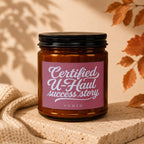 Amber glass candle with black lid, pink label reads "Certified U-Haul success story VOWED".