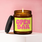 Amber jar candle with lit wick, lid off, reading "OPEN BAR LOADING... VOWED."