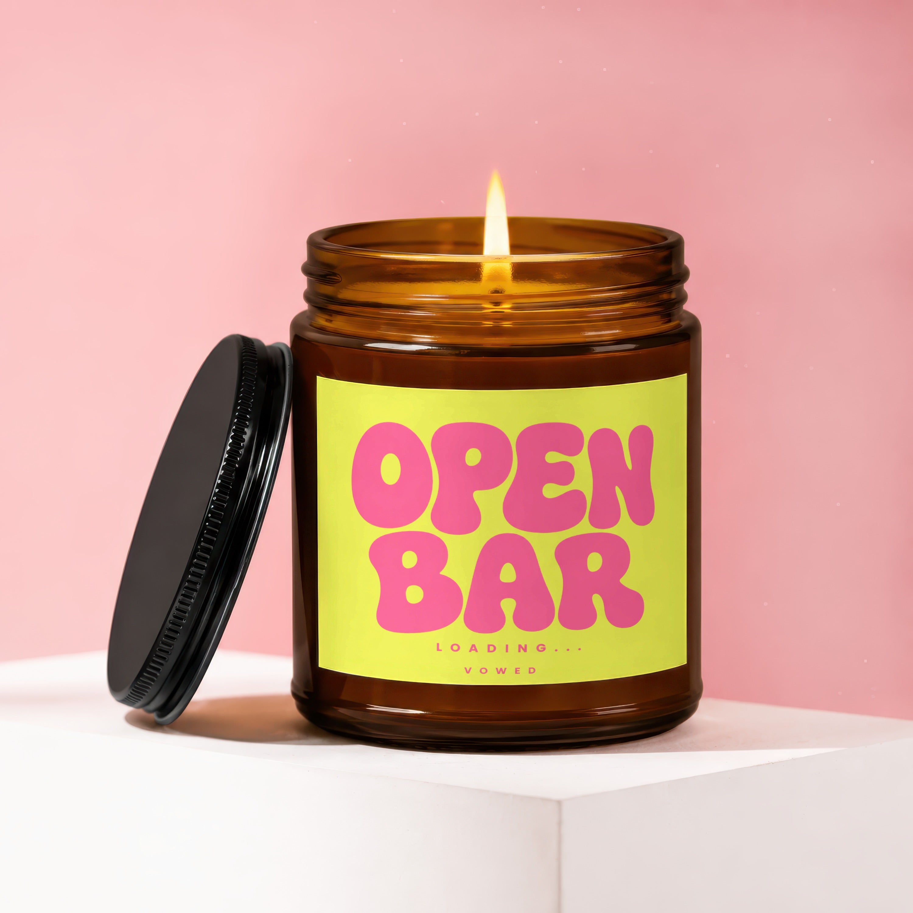 Amber jar candle with lit wick, lid off, reading "OPEN BAR LOADING... VOWED."