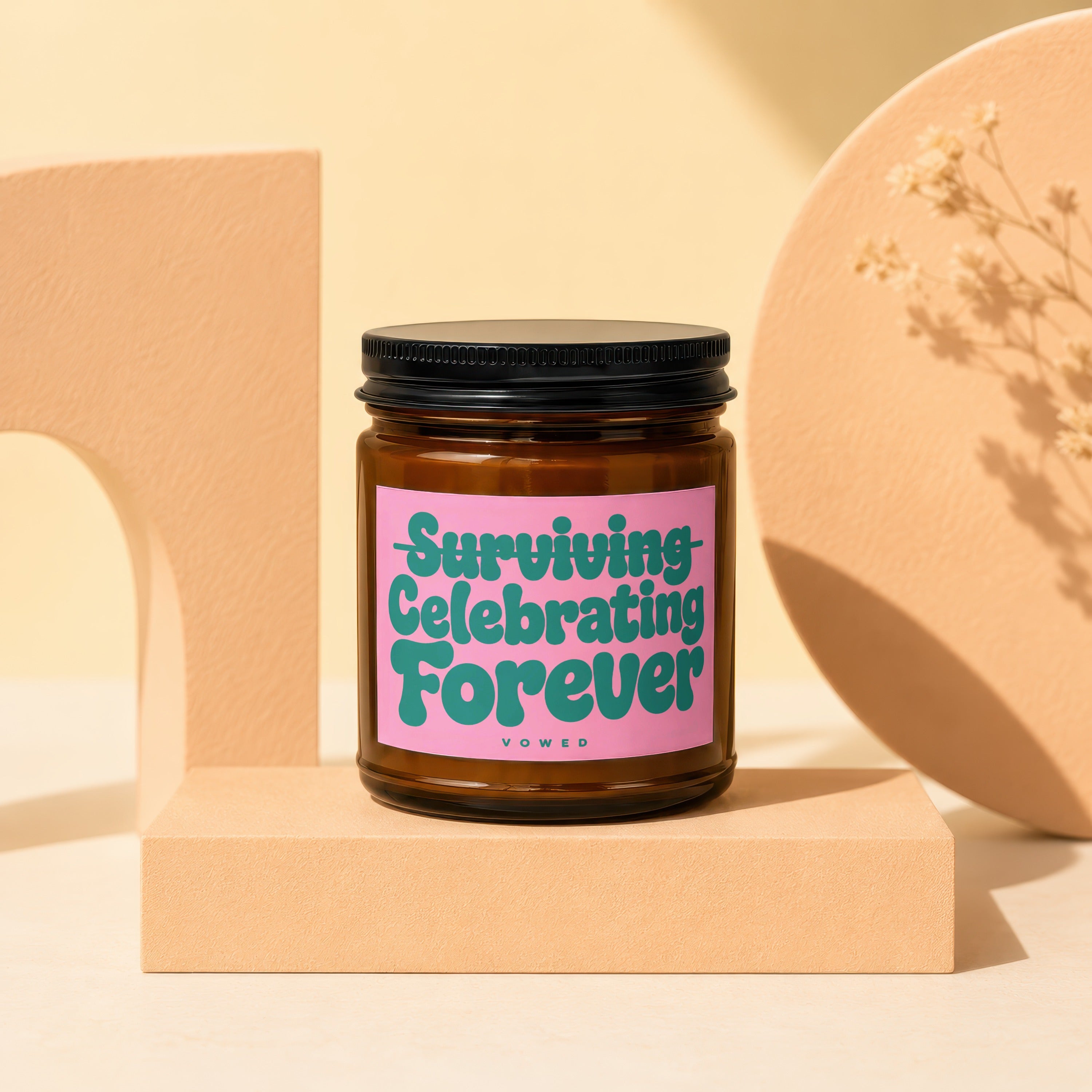Amber glass candle with pink label reading "Surviving Celebrating Forever" by VOWED.