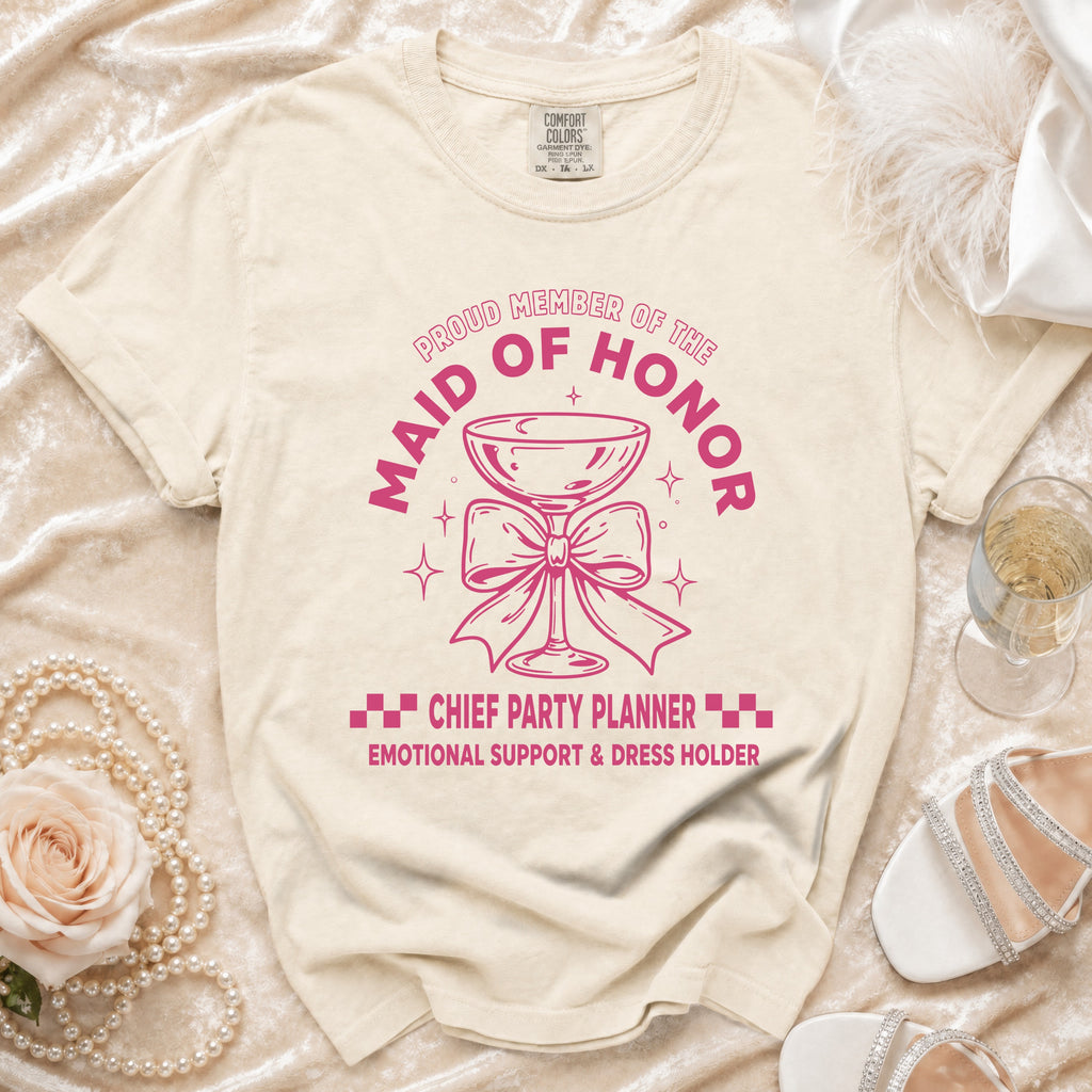 Light beige t-shirt with pink graphic of a champagne glass and text "Proud Member of the Maid of Honor Chief Party Planner."
