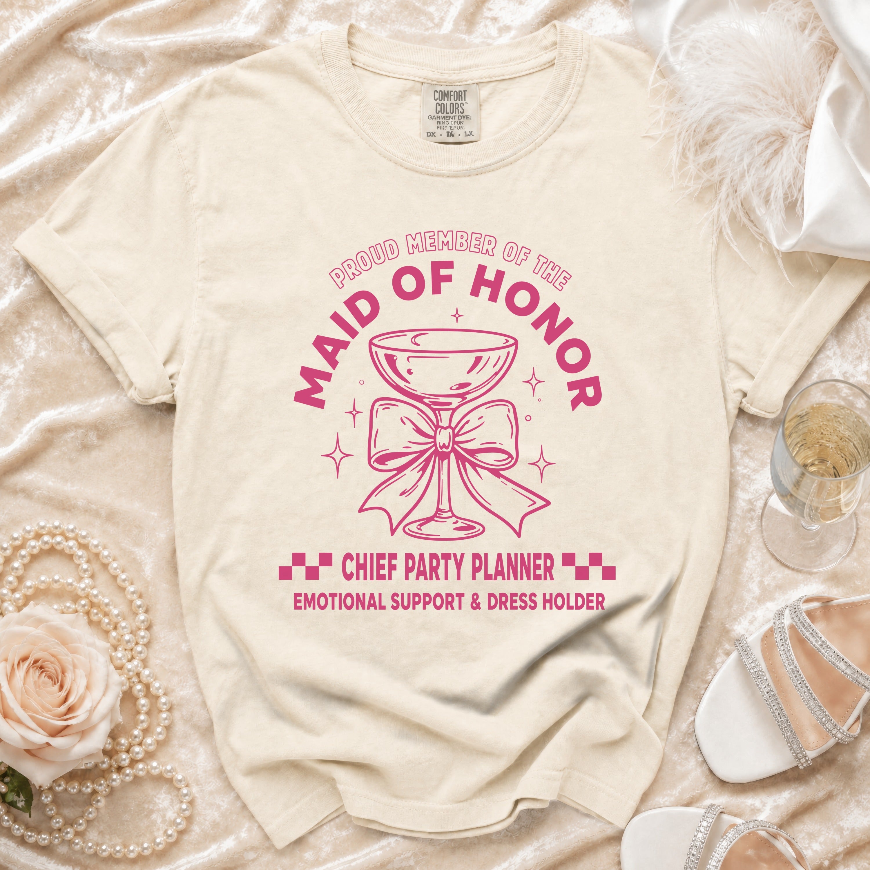 Light beige t-shirt with pink graphic of a champagne glass and text "Proud Member of the Maid of Honor Chief Party Planner."