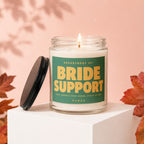 Candle with "Bride Support" label, open lid nearby, on white pedestal.