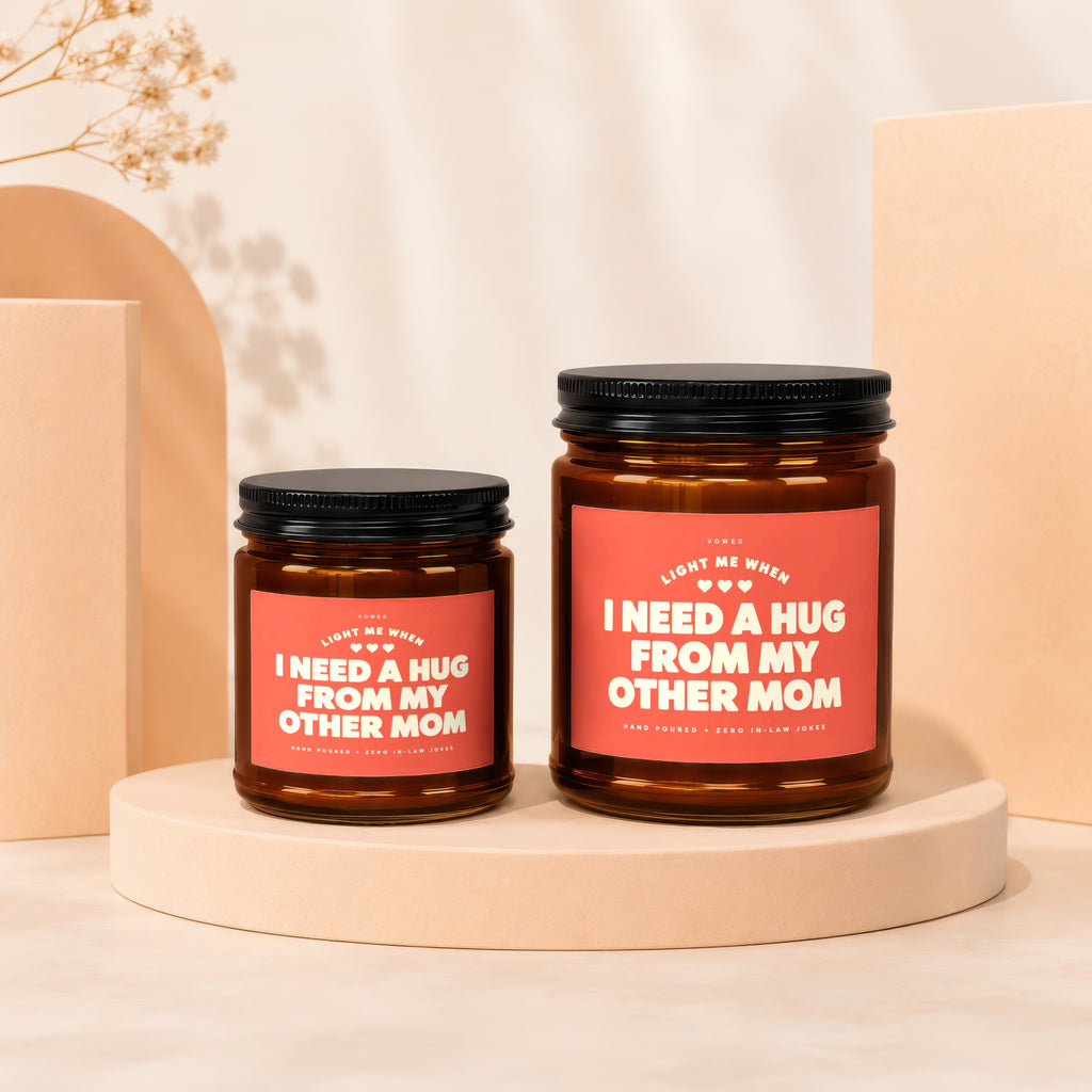 Two amber glass candles with black lids and coral labels saying "I need a hug from my other mom".
