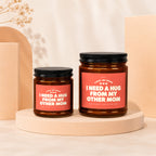 Two amber glass candles with black lids and coral labels saying "I need a hug from my other mom".