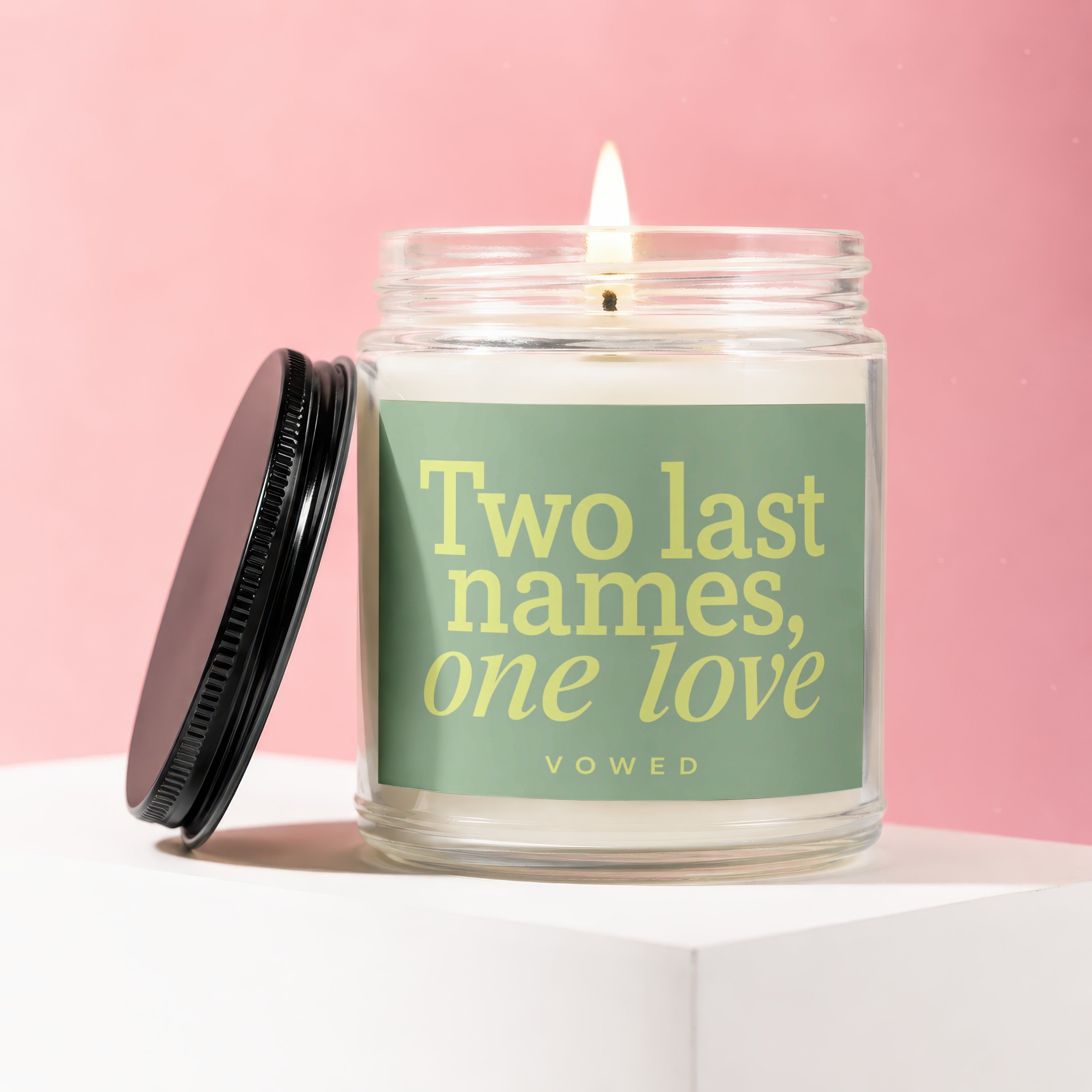 Lit candle in glass jar with "Two last names, one love" text and lid nearby.